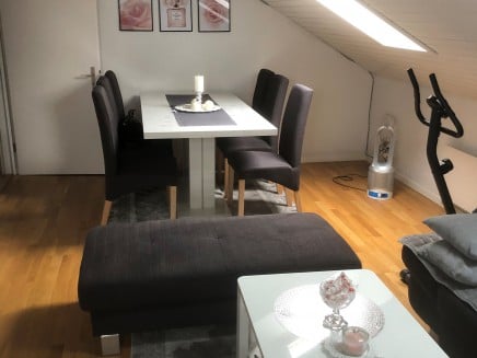Dining area with a white dining table and four chairs, a grey sofa, a small coffee table with a candle, and an exercise bike in the corner.