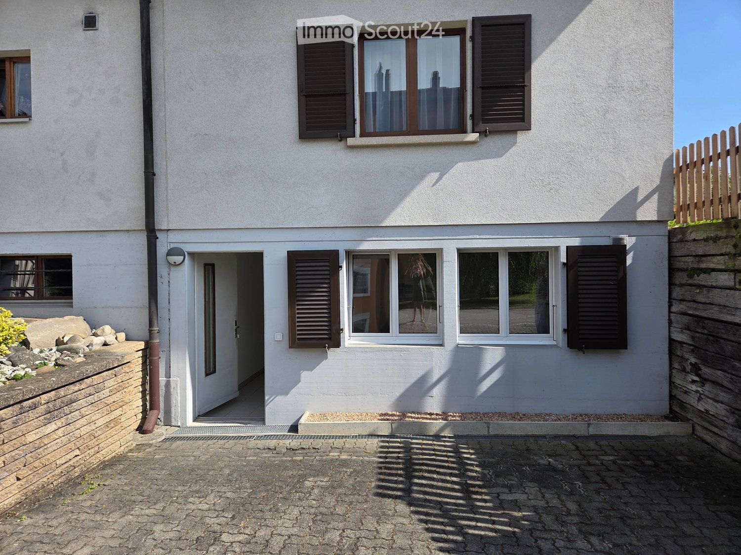 Two-story white building with multiple windows, shutters, and a raised entrance. The ground floor has a paved area with a small garden on the left.