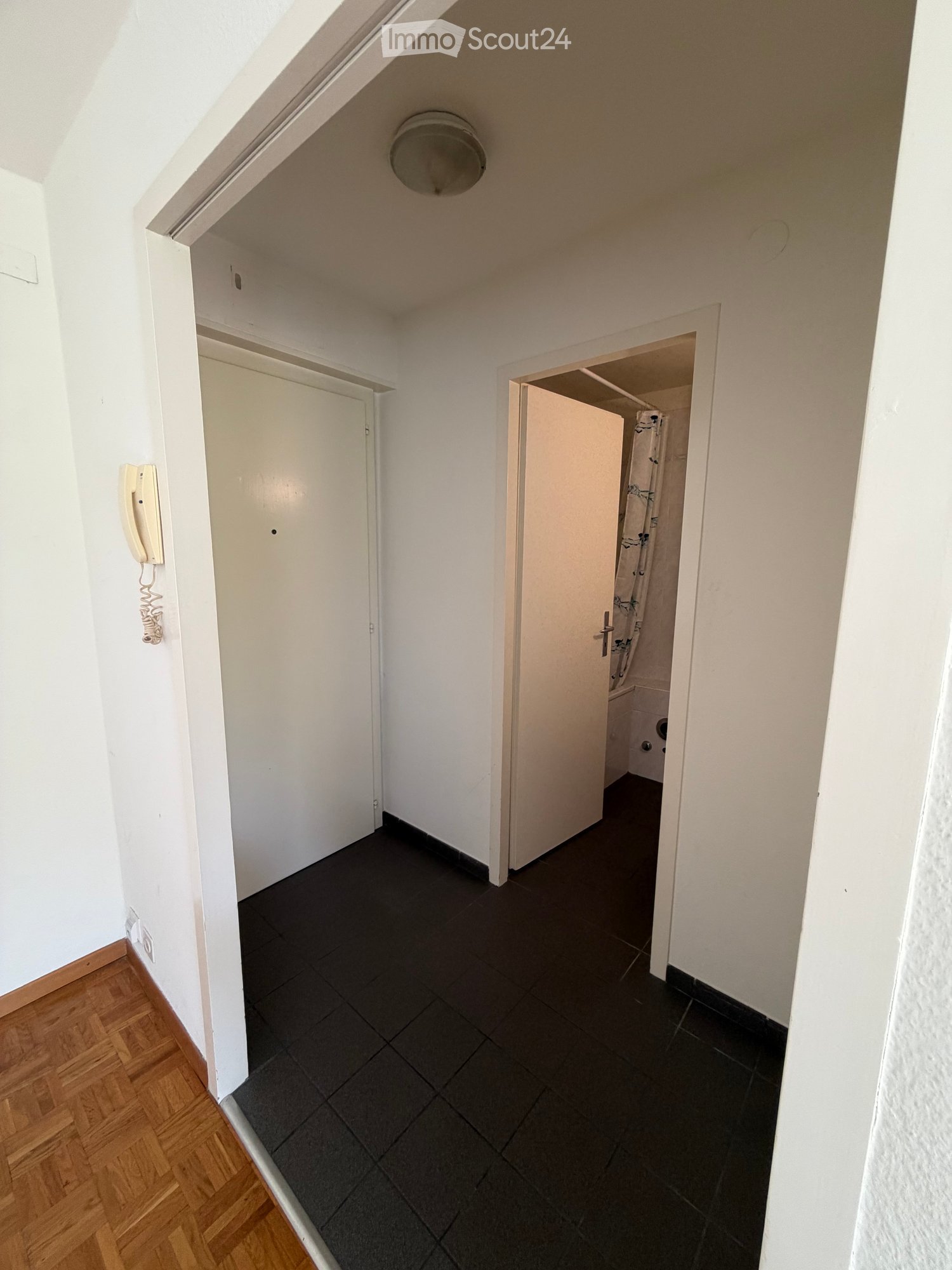 A narrow hallway with white walls, wooden flooring on one side, and dark tiled flooring on the other. Two doors are visible, one leading to a room and the other to a bathroom. A telephone is mounted on the wall.