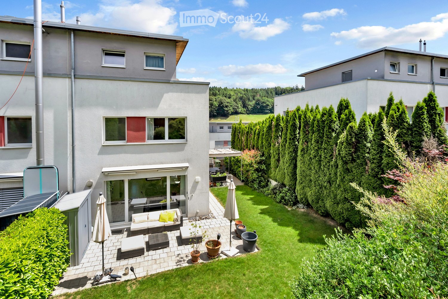 A modern two-story house with a spacious garden and patio area. The garden features neatly trimmed hedges, tall evergreen trees, and a well-maintained lawn. The patio includes outdoor furniture such as a sofa, ottomans, and a coffee table, along with pott