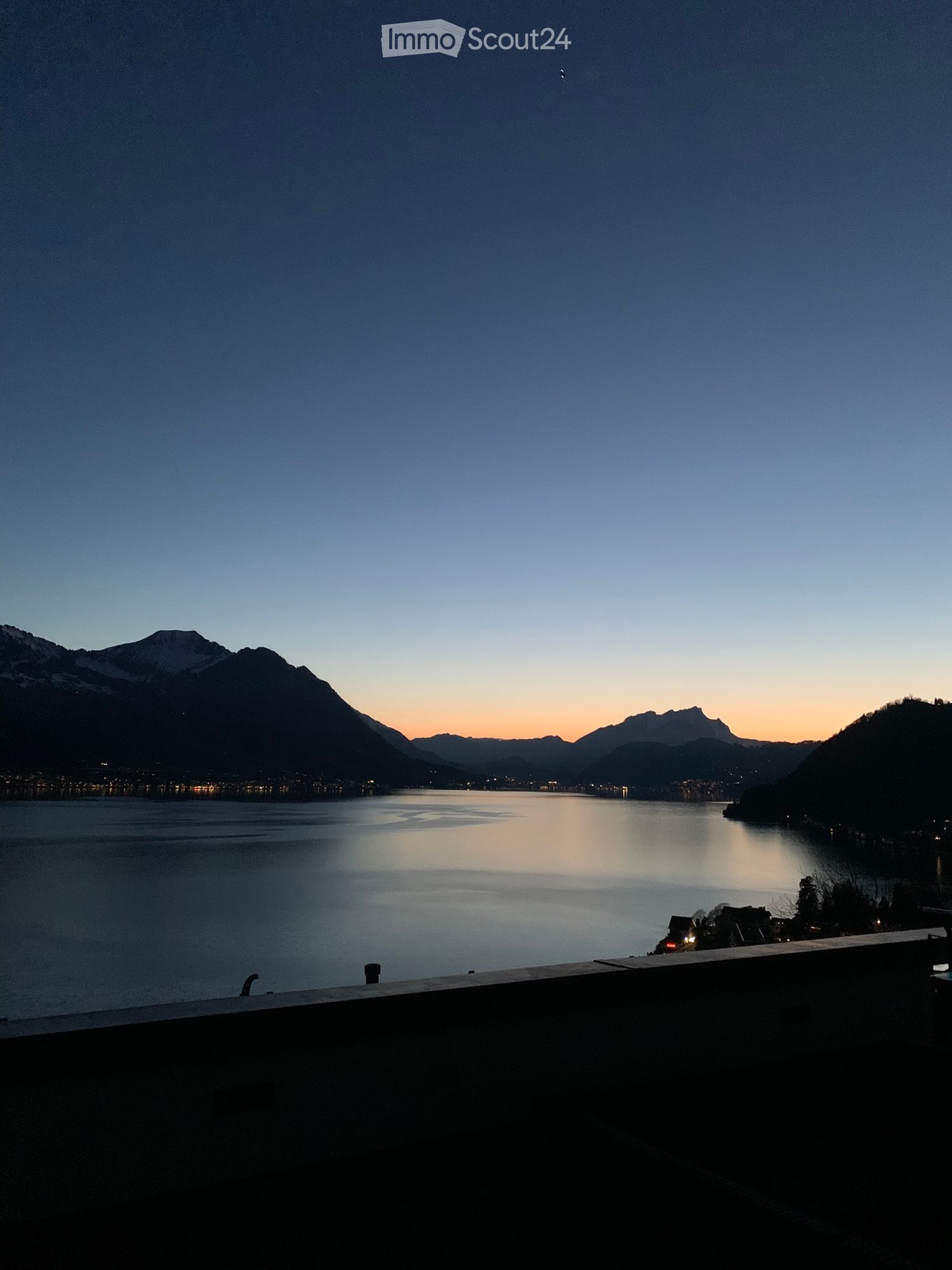 View from a balcony or terrace overlooking a serene lake with distant mountains during sunset.