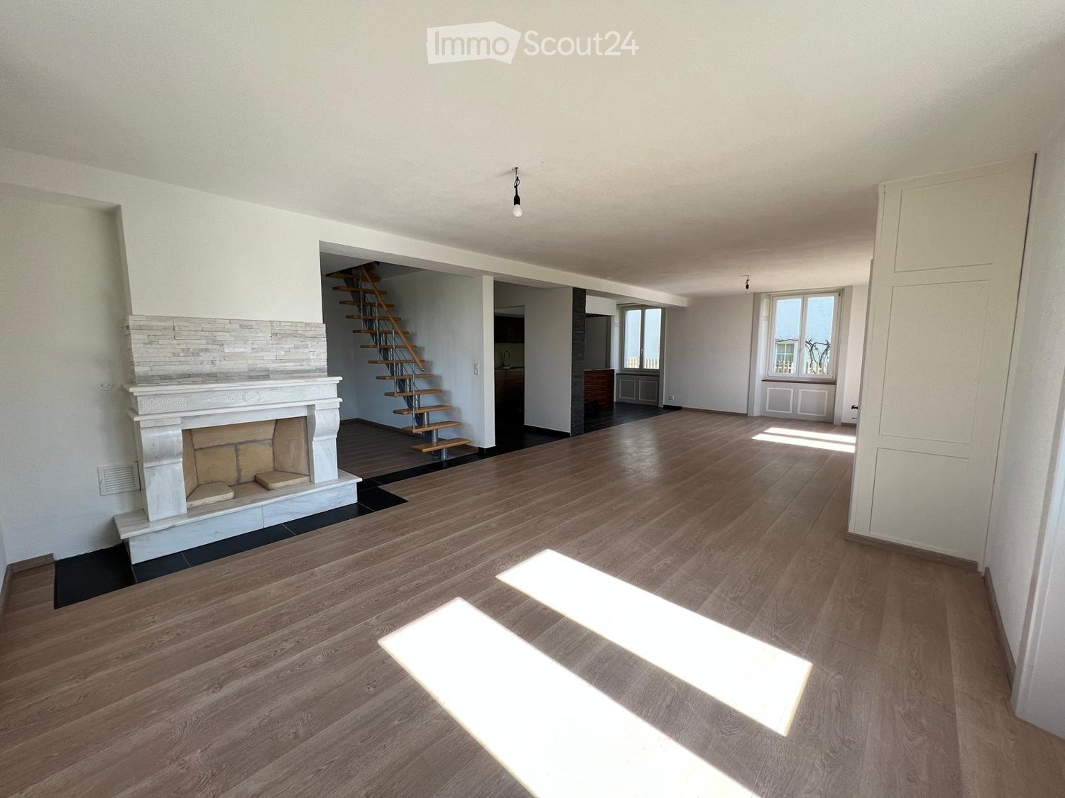 Empty room with wooden flooring, white walls, and a fireplace. A staircase leads to another level, and there are windows allowing natural light to enter.