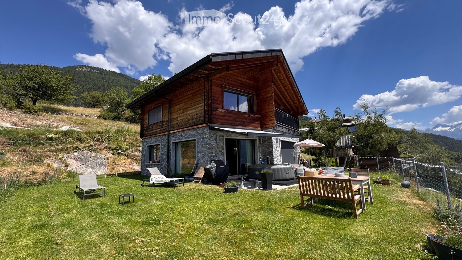 A two-story wooden house with stone base, large windows, and a spacious lawn with outdoor furniture including chairs, tables, and a bench.