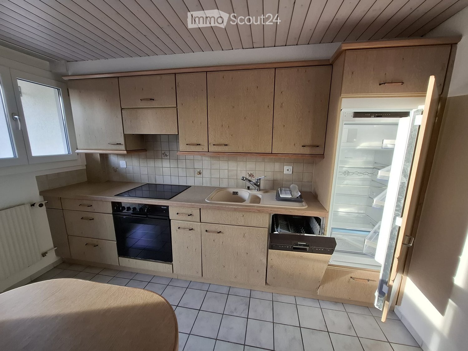 Modern kitchen with light wooden cabinets, tiled walls, and appliances including a sink, oven, dishwasher, and refrigerator. A round wooden table is partially visible in the foreground.