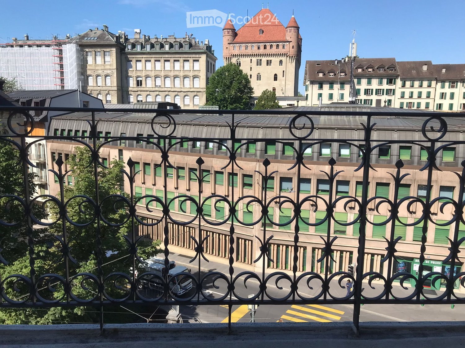 Balcony with ornate metal railing overlooking a cityscape with historic buildings, modern structures, and a visible street below.