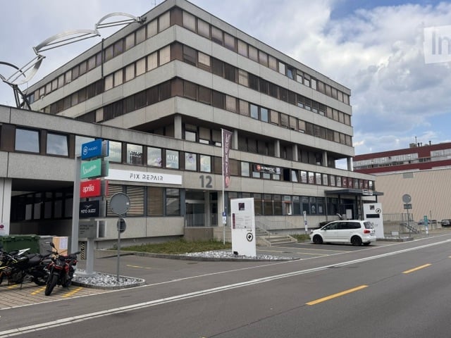 Multi-story commercial building with a modern facade, signage for various businesses, and a parking area with motorcycles and a car.