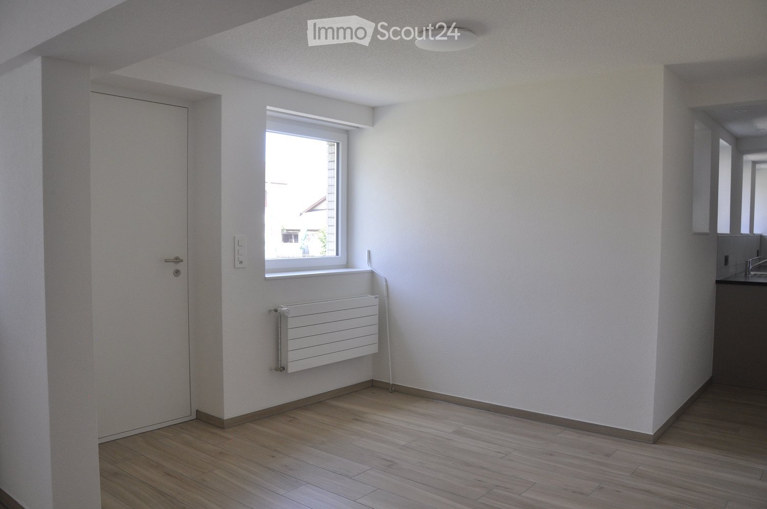 Empty room with wooden flooring, white walls, a window, a radiator, and a door. No furniture or equipment is present.