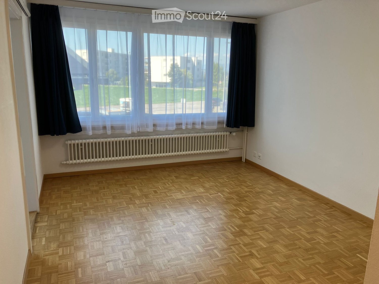 Empty room with wooden herringbone flooring, white walls, large window with sheer curtains, radiator, and visible electrical outlet.