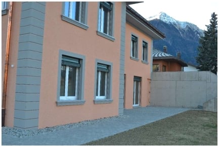 A two-story residential building with a peach-colored facade, grey window frames, and a small driveway leading to a garage entrance. The background shows a mountainous landscape.