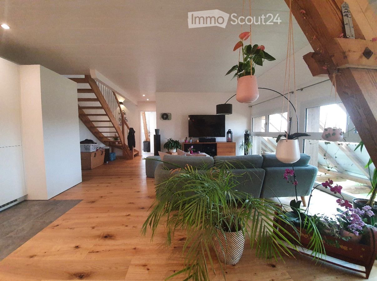 Modern living room with wooden flooring, grey sofa, coffee table, television, floor lamp, and several potted plants. A wooden staircase leads to an upper level.
