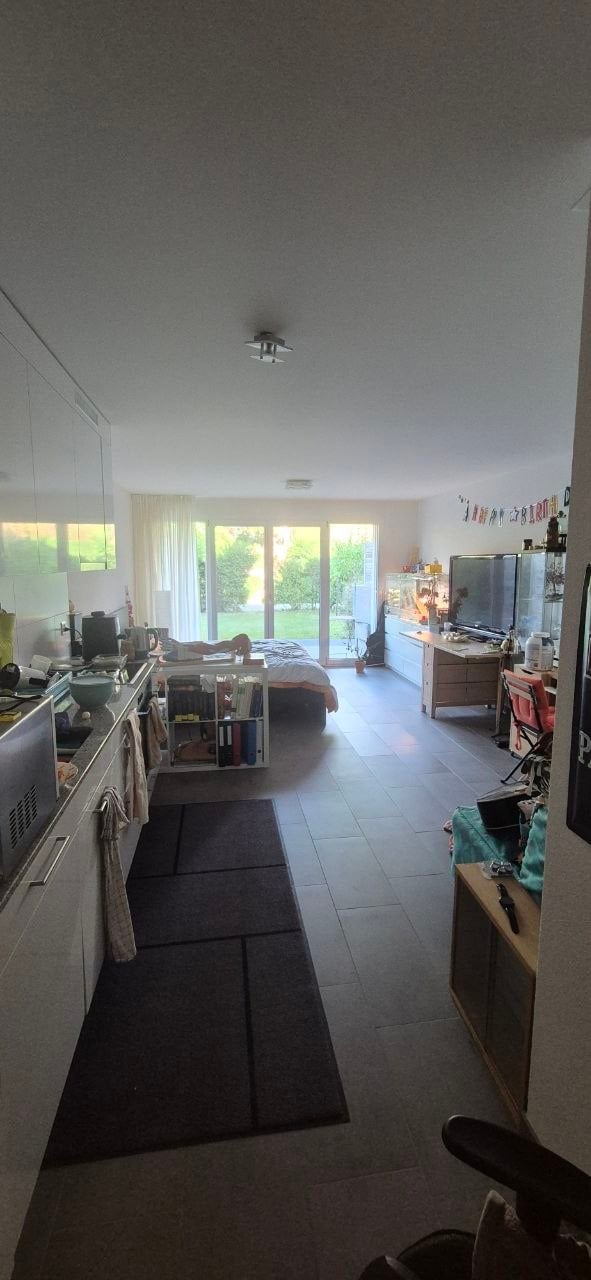 Open-plan living and kitchen area with modern kitchen cabinets, a sink, fridge, microwave, oven, and a small bed in the living area. There is a laptop on a table, a screen, a bookshelf, and a curtain covering a large window.
