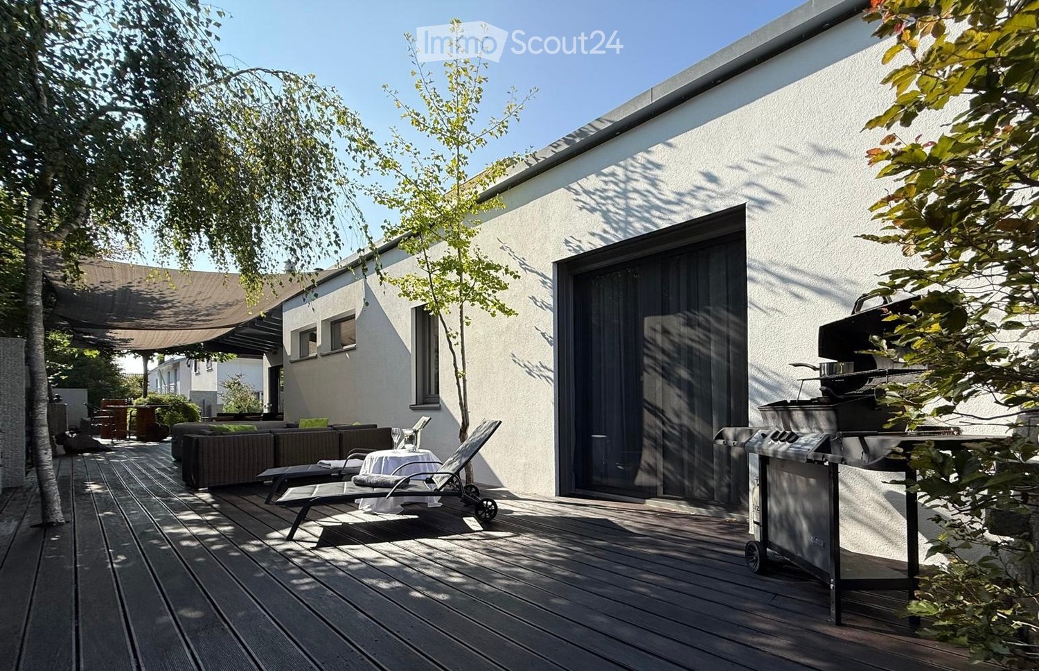 Wooden deck area with a modern house in the background, featuring a grill, lounge chairs, a small table, and a sofa.