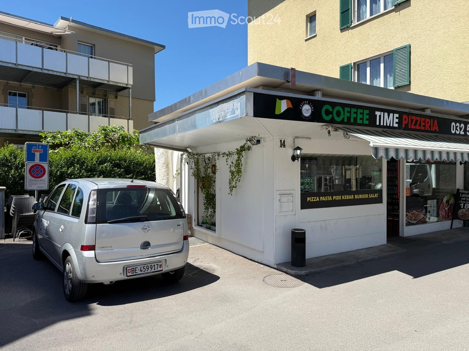 Small, modern building with a sign reading "COFFEE TIME PIZZERIA" and a menu board listing pizza, pasta, pide, kebab, burger, and salad. A silver car is parked nearby, and there is a balcony on the adjacent building.