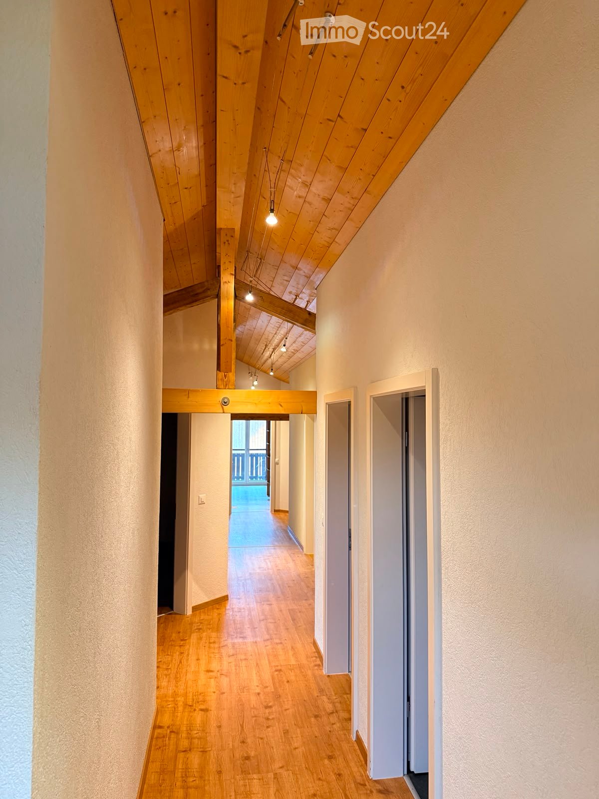 Wooden hallway with exposed wooden beams and recessed lighting, leading to an open area with a view of a balcony.