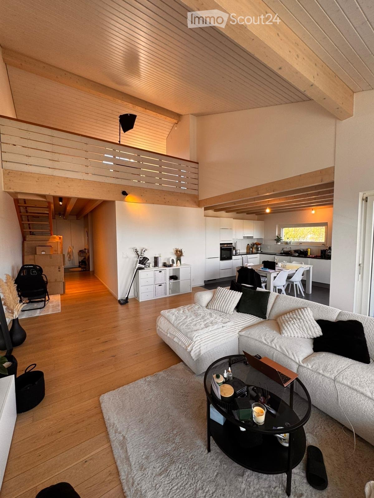 Modern living room with a white sectional sofa, black coffee table, laptop, dining area with white chairs and table, kitchen area with white cabinets and appliances, and a staircase leading to an upper level.