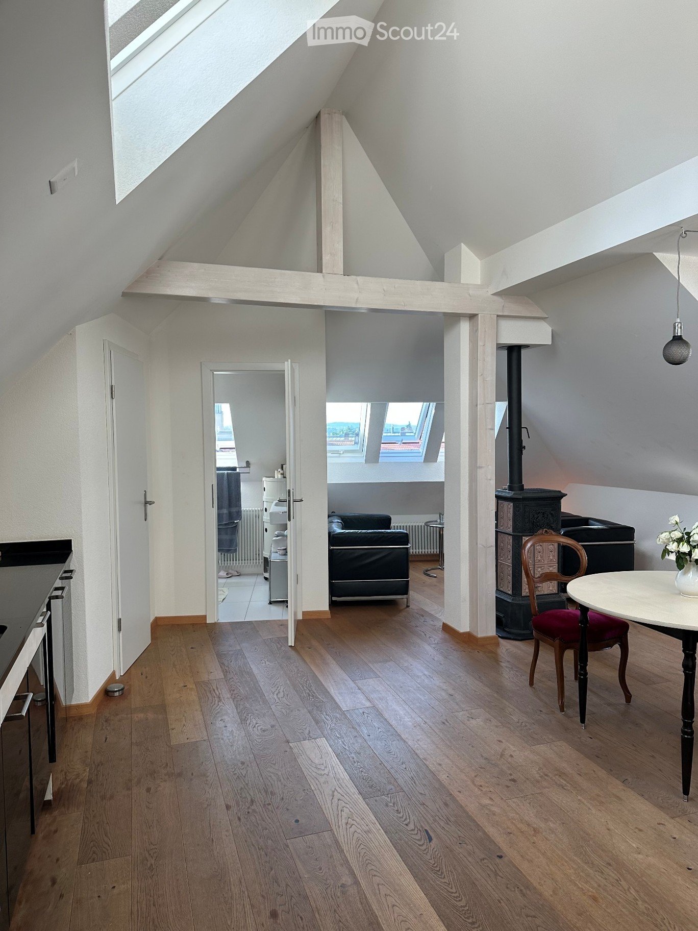 Bright, open-plan attic space with wooden flooring, exposed wooden beams, and skylights. Features a round table with a chair, a sofa in an adjacent room, and a radiator.