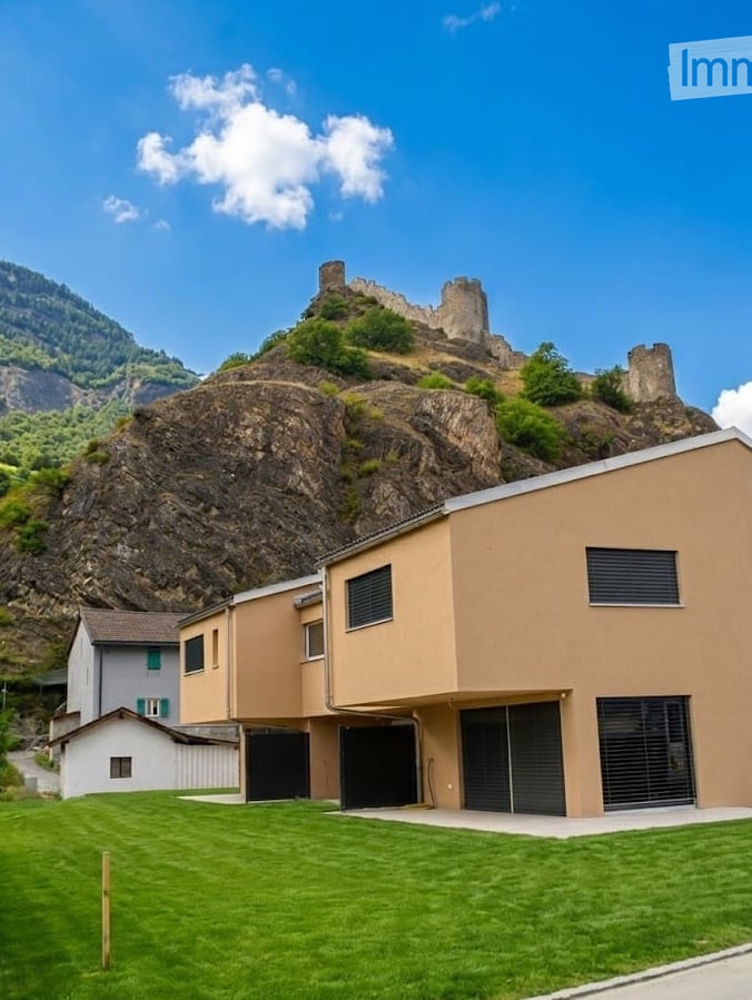 Modern two-story building with beige facade, large garage doors, and a well-maintained lawn, situated in front of a rocky hill with a castle-like structure on top.