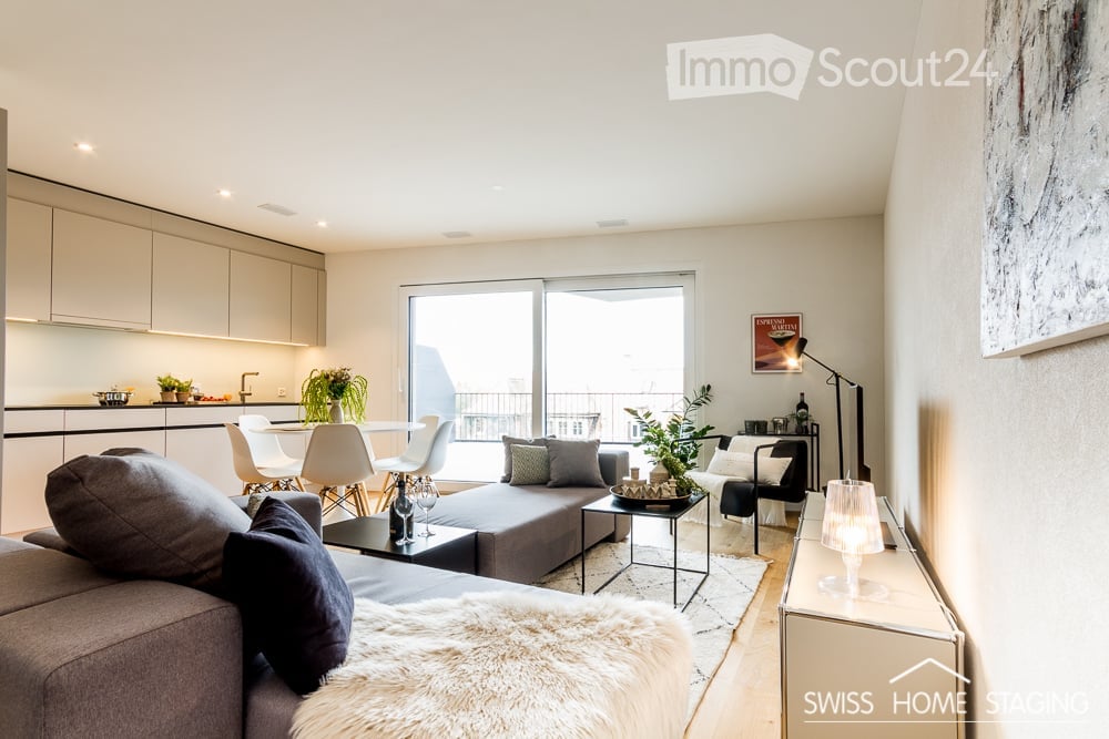Modern open-plan living and dining area with a grey sofa, coffee table, armchair, ottoman, floor lamp, dining table with chairs, kitchen cabinet, faucet, plants, picture frame, bottle, and wine glass.