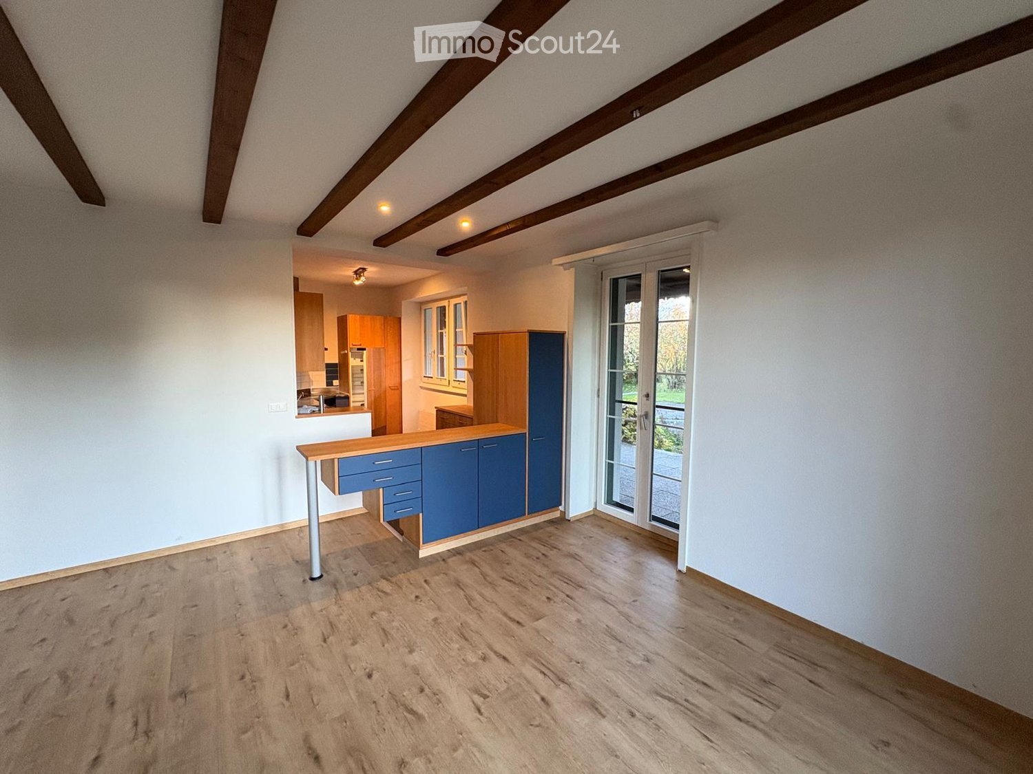 Bright, empty room with wooden flooring, white walls, and exposed wooden beams on the ceiling. Features a sliding glass door leading to an outdoor area and a small kitchen area visible in the background.