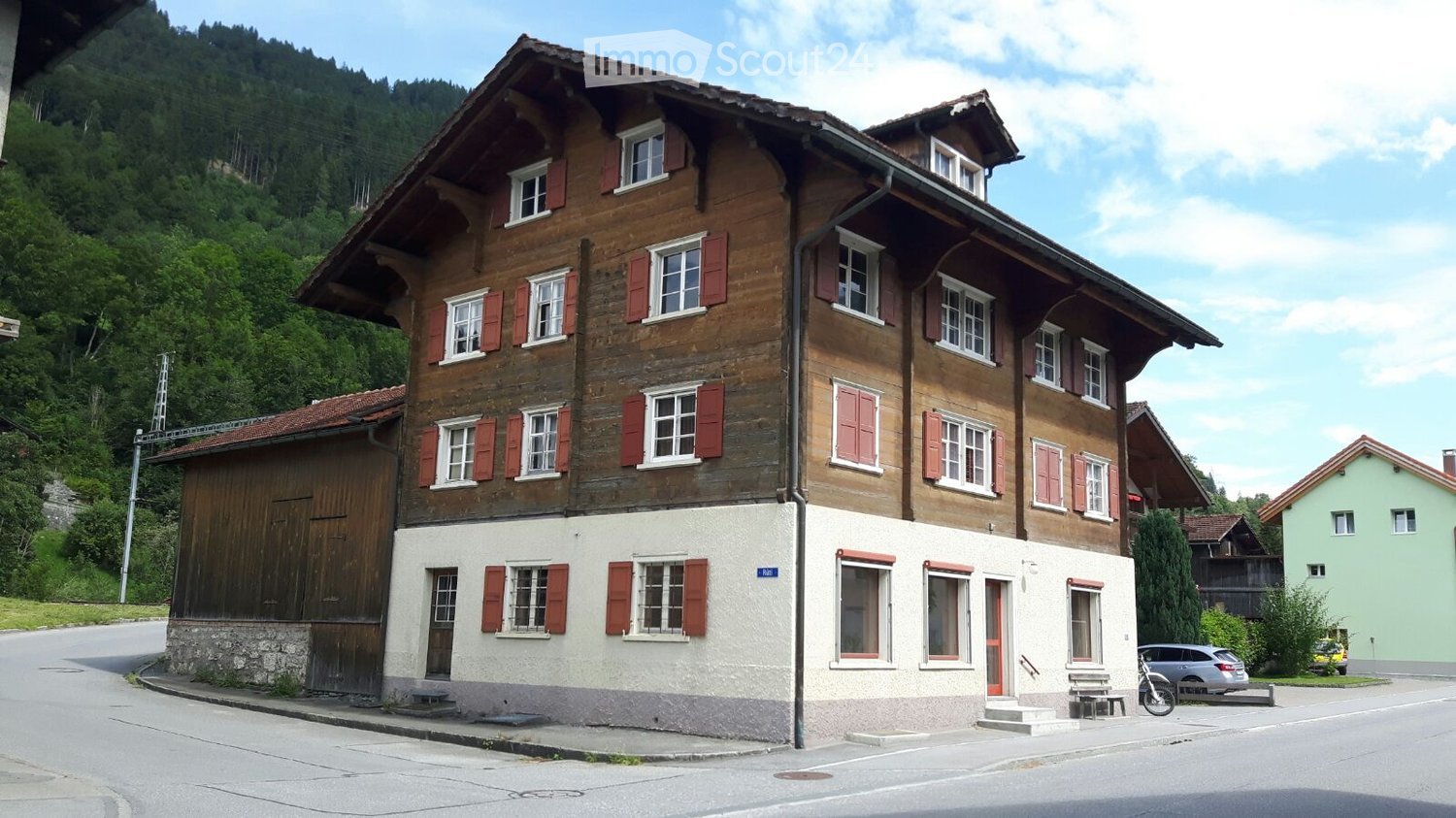 Traditional wooden house with red shutters, multiple windows, and a stone base, situated in a scenic, mountainous area.