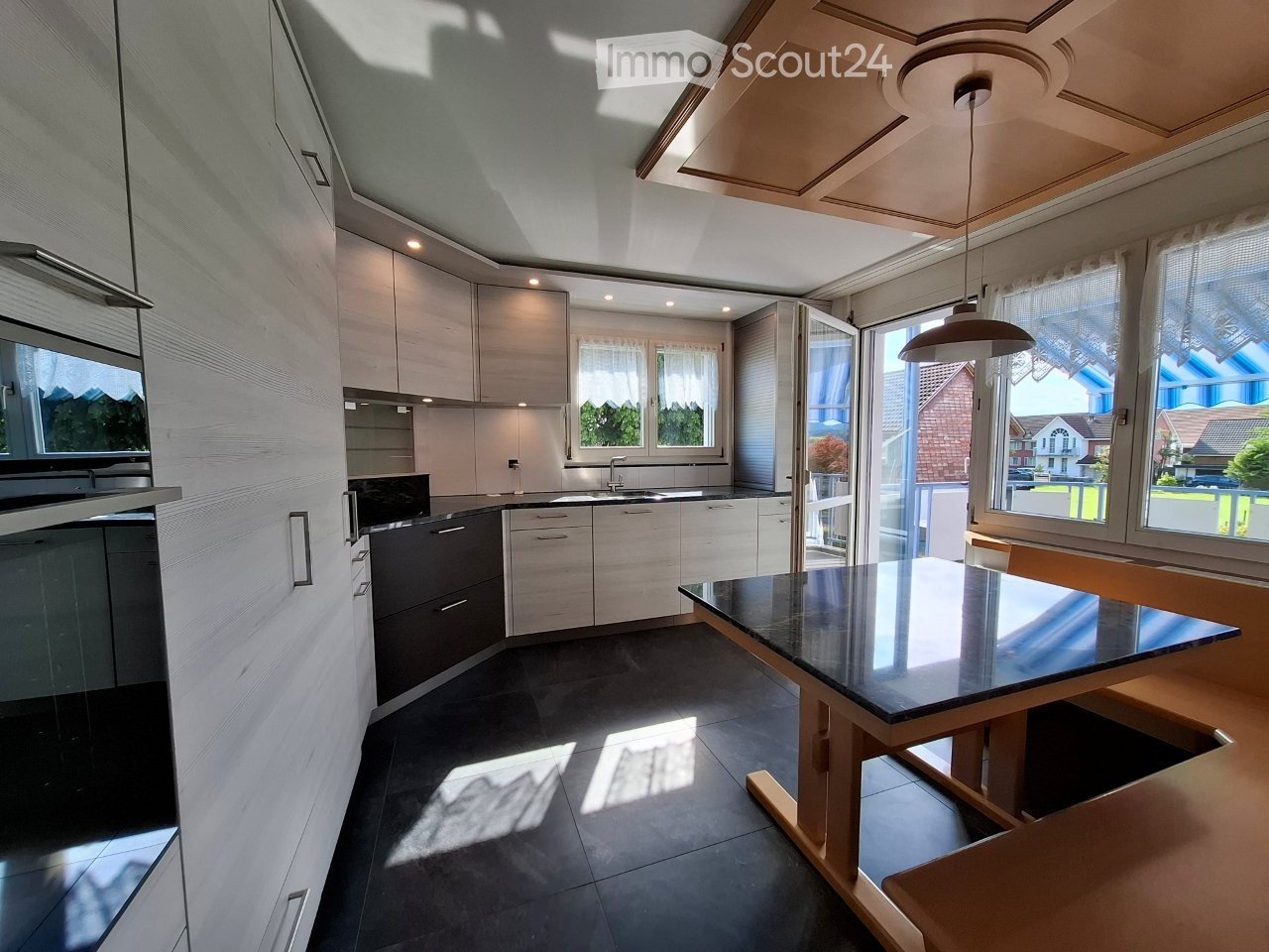 Modern kitchen with light-colored cabinets, a central island, and a dining area with a black countertop table and wooden chairs. The space features large windows and a pendant light hanging from the ceiling.