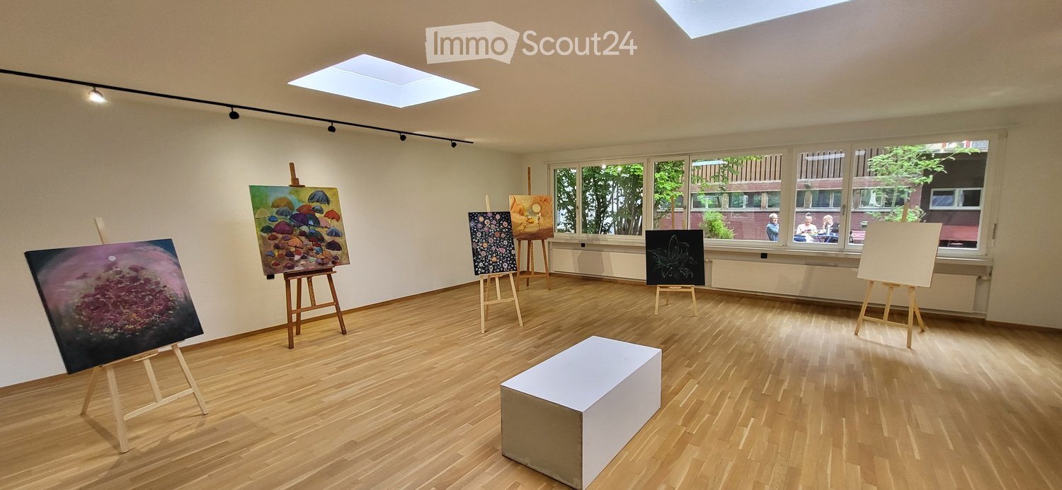 An art studio with wooden floors, large windows, and several easels displaying paintings. A white stool is placed in the center of the room.