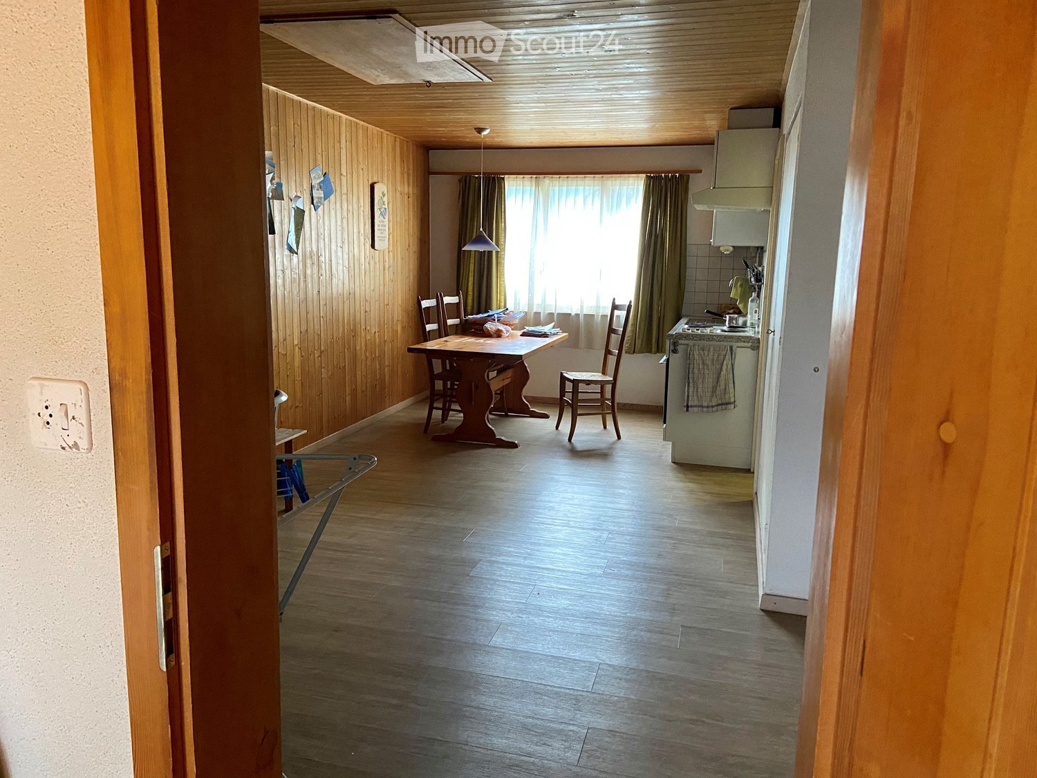 A furnished room with a wooden table and chairs, a kitchen area with a stove and fridge, and wooden walls and flooring.
