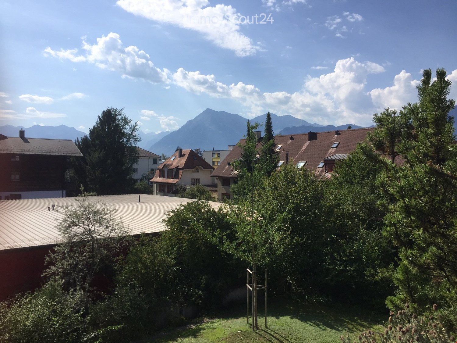 A serene garden area with lush greenery, including trees and shrubs, surrounded by residential houses and a scenic mountain backdrop under a partly cloudy sky.