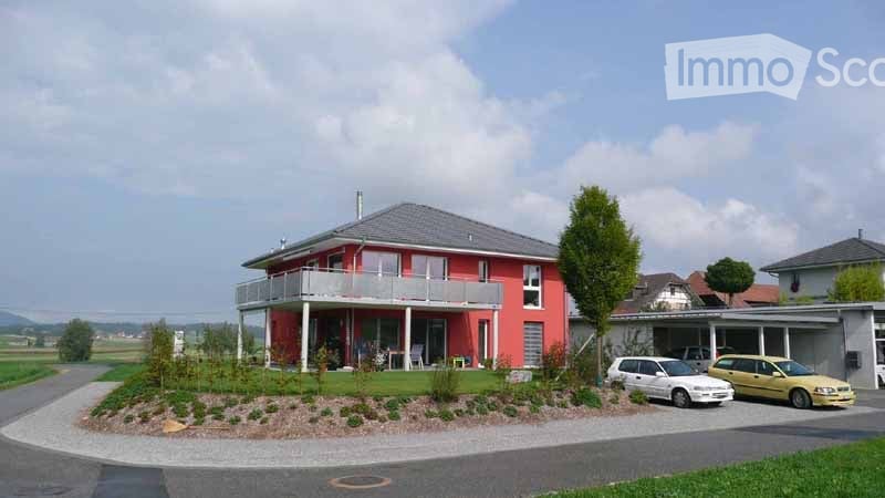 Red single-story house with a grey roof, white-framed windows, and a balcony on the front. There is a small garden area with shrubs and a driveway with two parked cars. A garage is visible to the right.