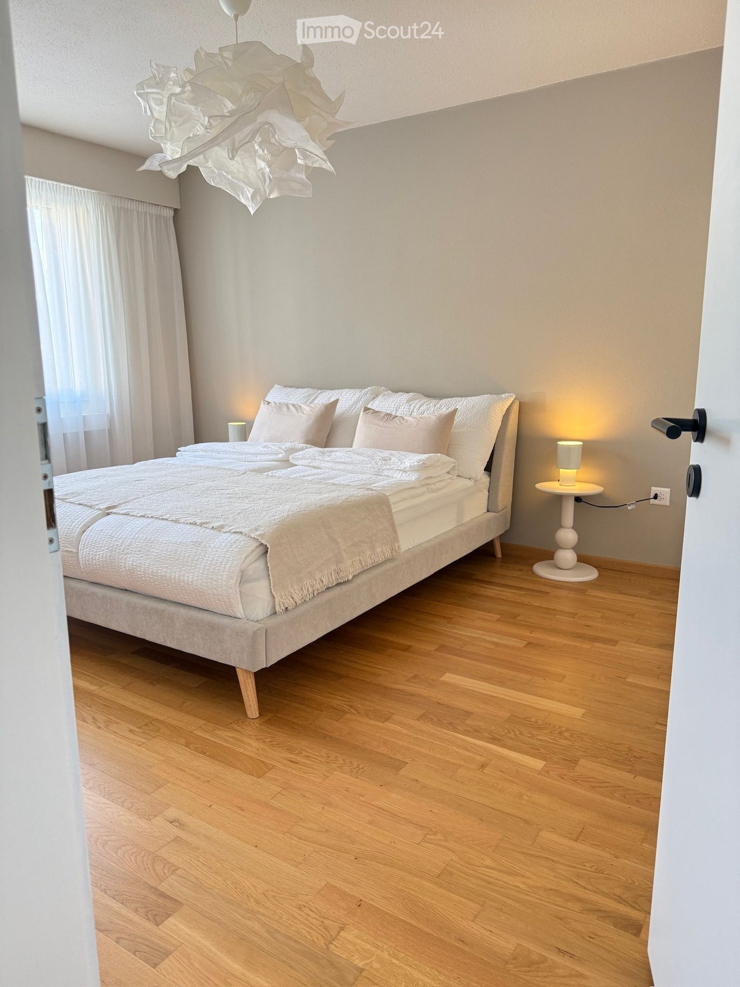 Modern bedroom with a large bed, white bedding, wooden floor, nightstand with lamp, and sheer curtains.