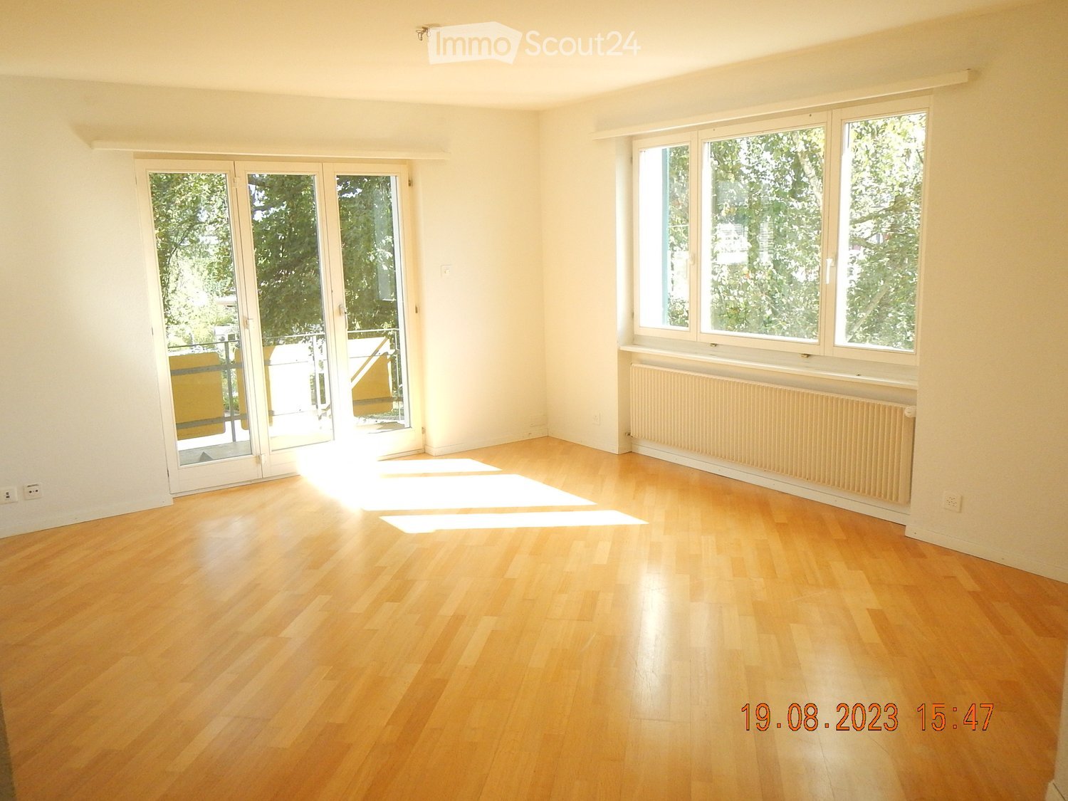 Empty room with light wooden flooring, large windows, and sliding glass doors leading to a balcony.