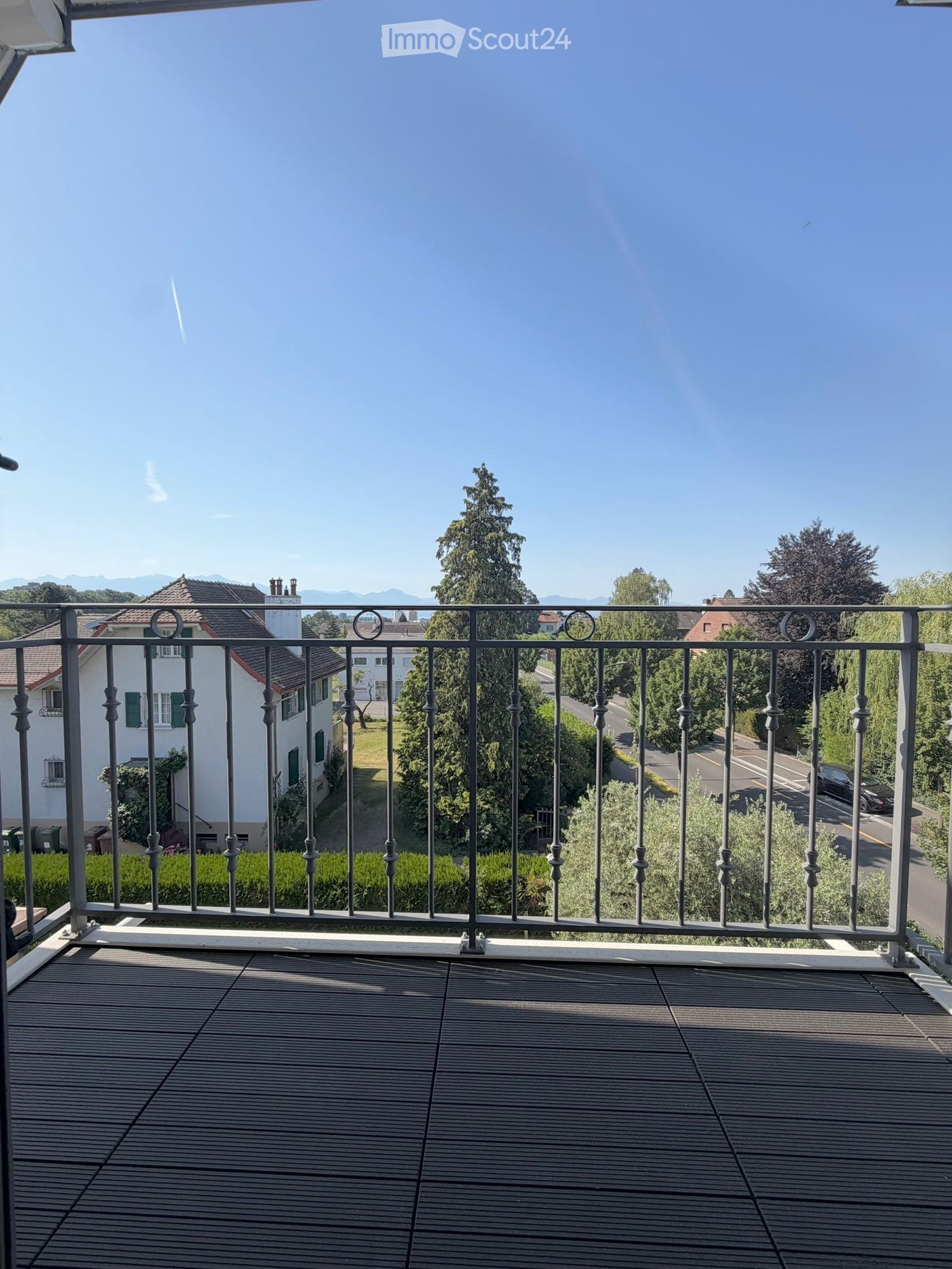 Balcony with wooden decking and metal railing, overlooking a residential area with houses, trees, and a clear blue sky.