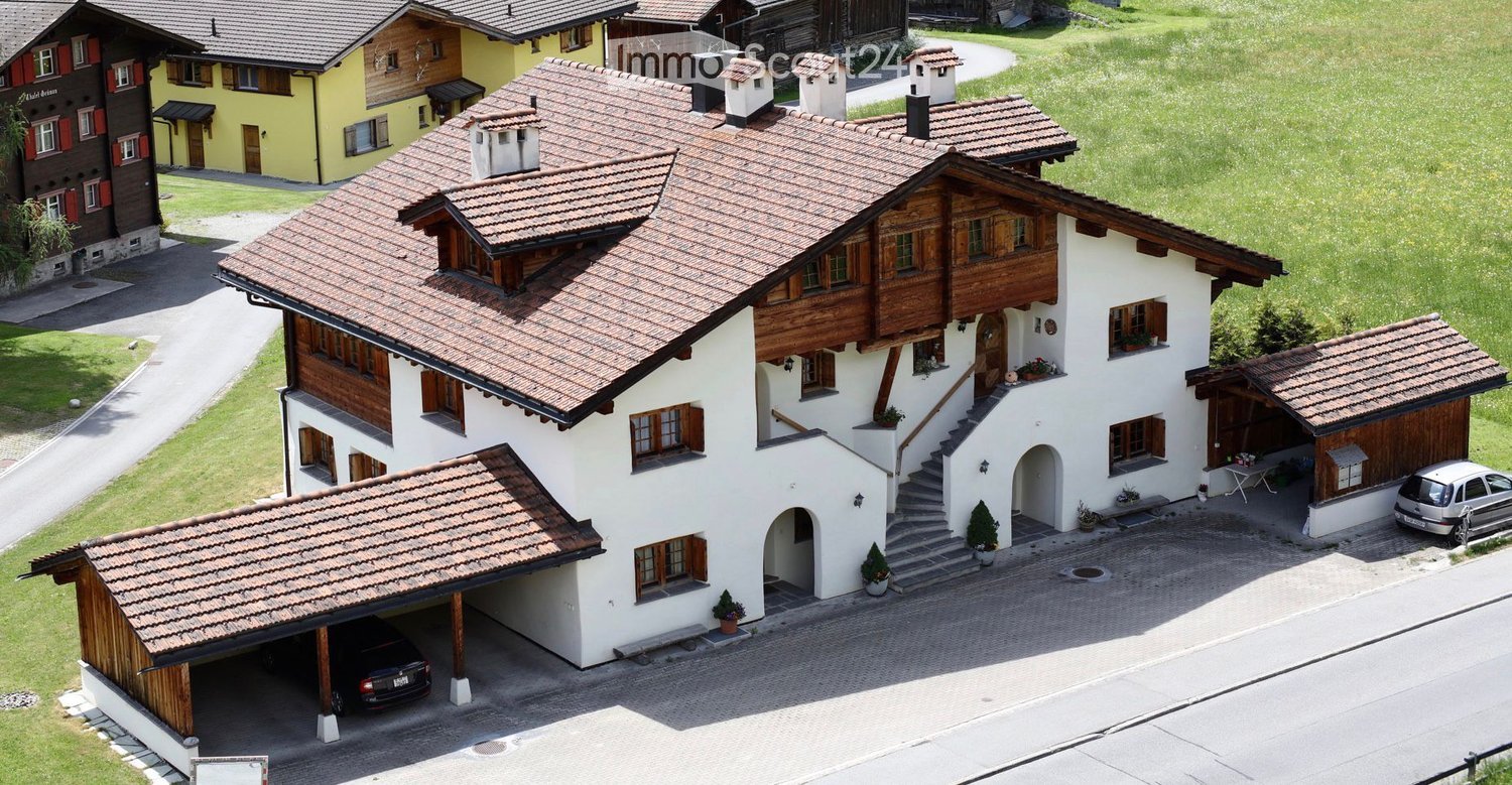 A large, two-story house with a tiled roof, white walls, and wooden accents. Features include a garage, a driveway, and surrounding greenery.