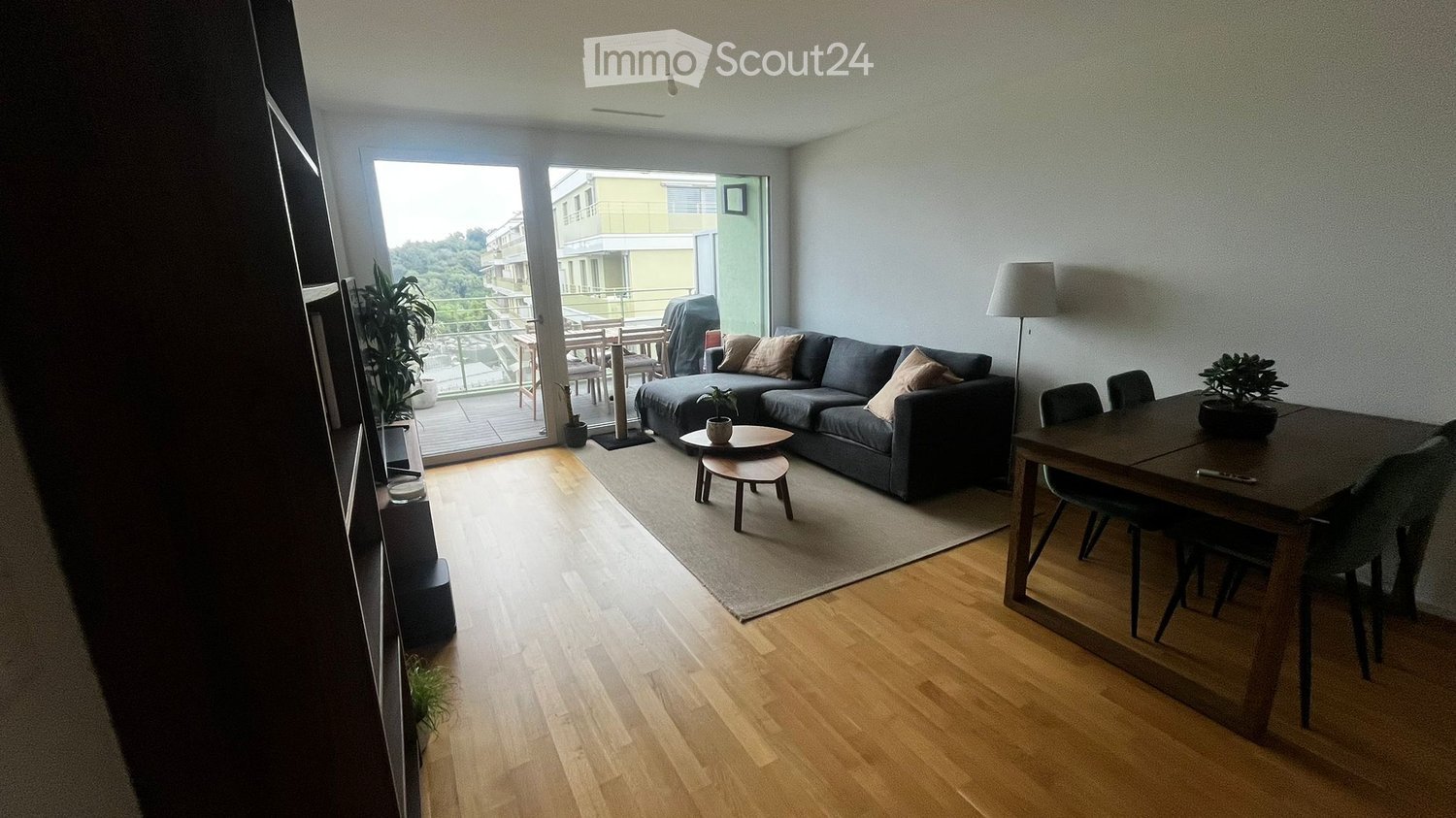 Modern living room with wooden flooring, grey sofa, coffee table, dining table, and balcony access.