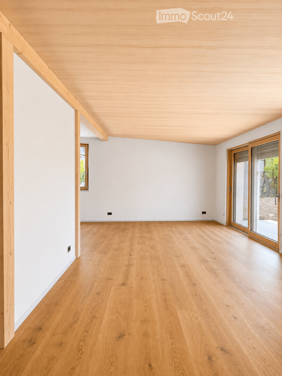 Empty room with light wooden flooring, white walls, and a wooden ceiling. Features a large sliding glass door leading outside and a smaller window on the left side.