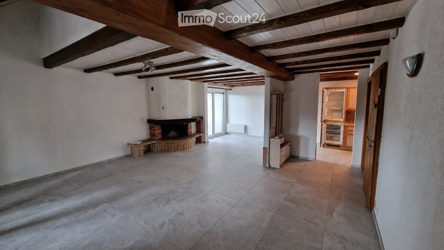 Empty hallway with exposed wooden beams, tiled floor, and a brick fireplace on the left side. A doorway leads to a kitchen area with visible cabinets and a refrigerator.