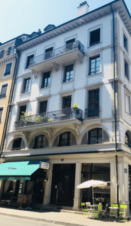 Multi-story building with white facade, multiple balconies, and a small outdoor seating area with tables and chairs.