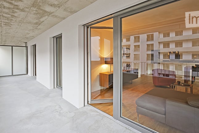 Modern balcony with large glass sliding doors opening to a furnished room, featuring a view of a contemporary building exterior.