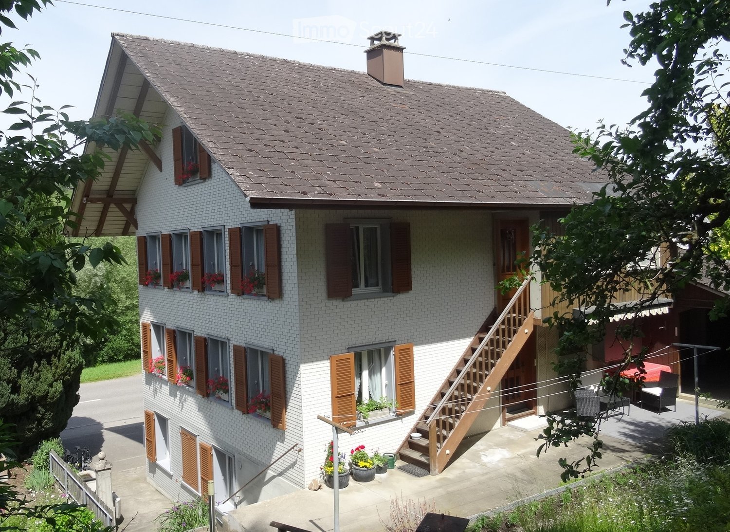 Two-story house with white brick exterior, brown shutters, and a wooden staircase leading to a raised ground floor. The house features multiple windows with flower boxes, a chimney, and a small garden area with potted plants and outdoor furniture.