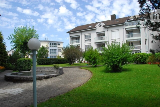 Well-maintained garden area with a circular stone fountain, neatly trimmed hedges, and a paved walkway leading to a multi-story residential building with balconies.