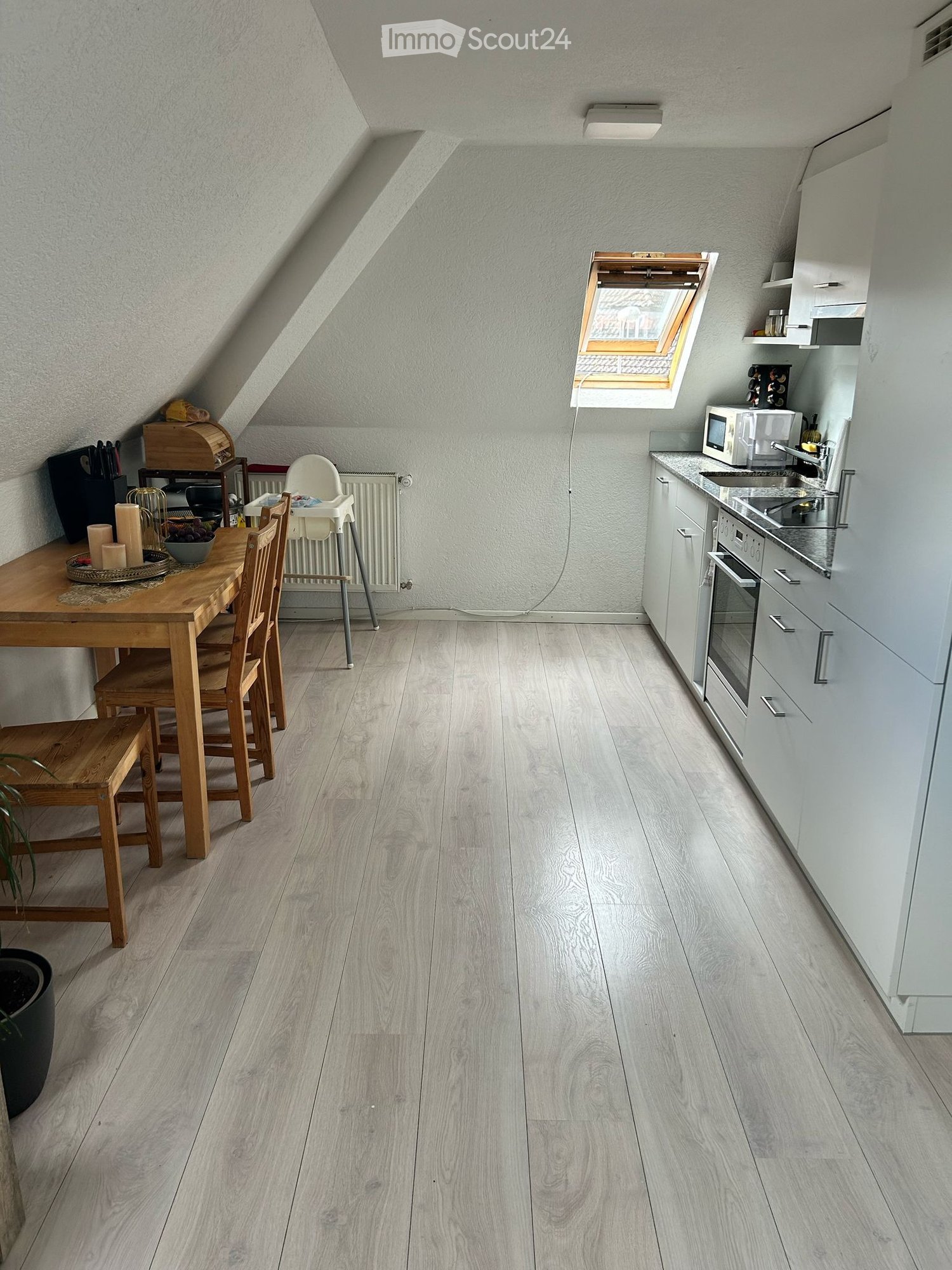 Modern kitchen and dining area with light wood flooring, white cabinets, and a wooden dining table with chairs. Features include a microwave, oven, sink, and a small window with blinds.
