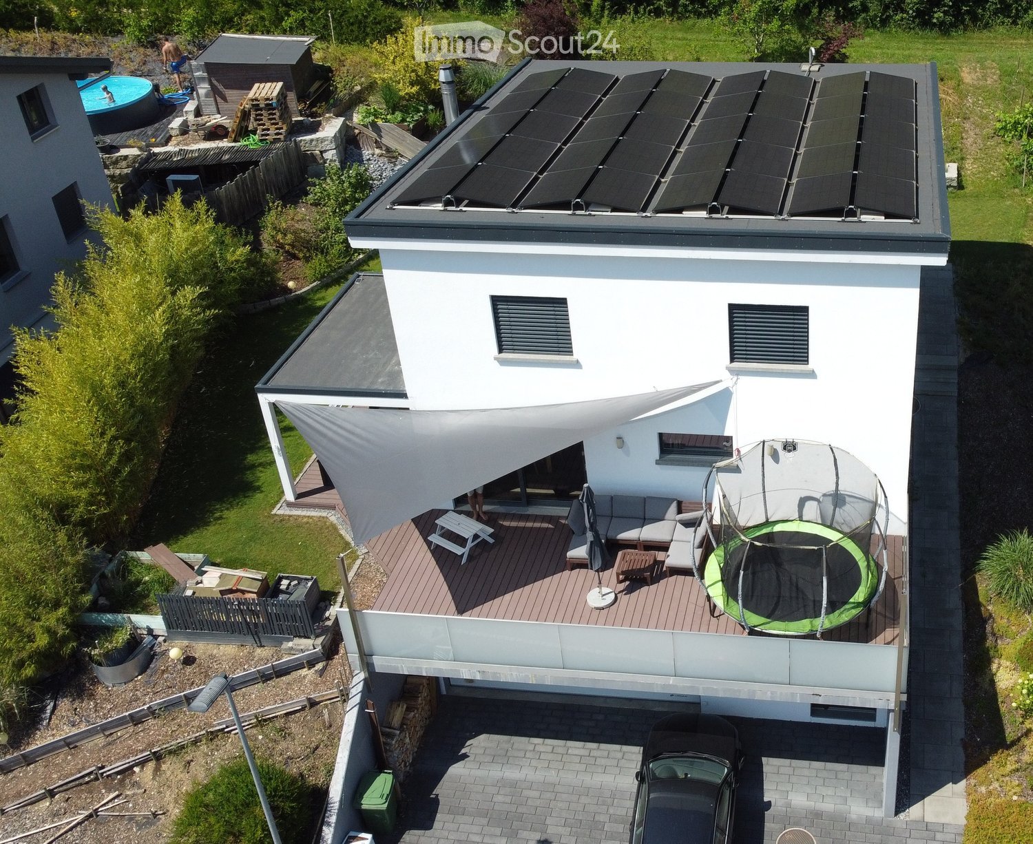 Modern two-story house with white exterior, solar panels on the roof, and a spacious outdoor deck with seating, umbrella, and trampoline. The surrounding area includes a swimming pool, garden, and additional structures.