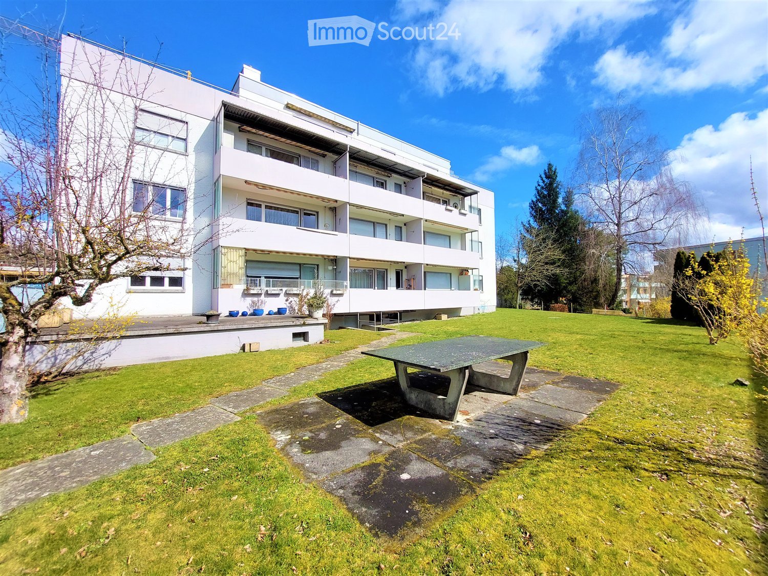 Modern white multi-story building with balconies and large windows, surrounded by a well-maintained lawn and trees.