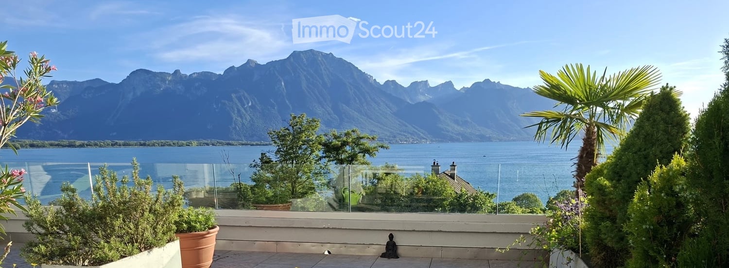 Balcony with glass railing overlooking a lake and mountain range, decorated with potted plants and a small statue.