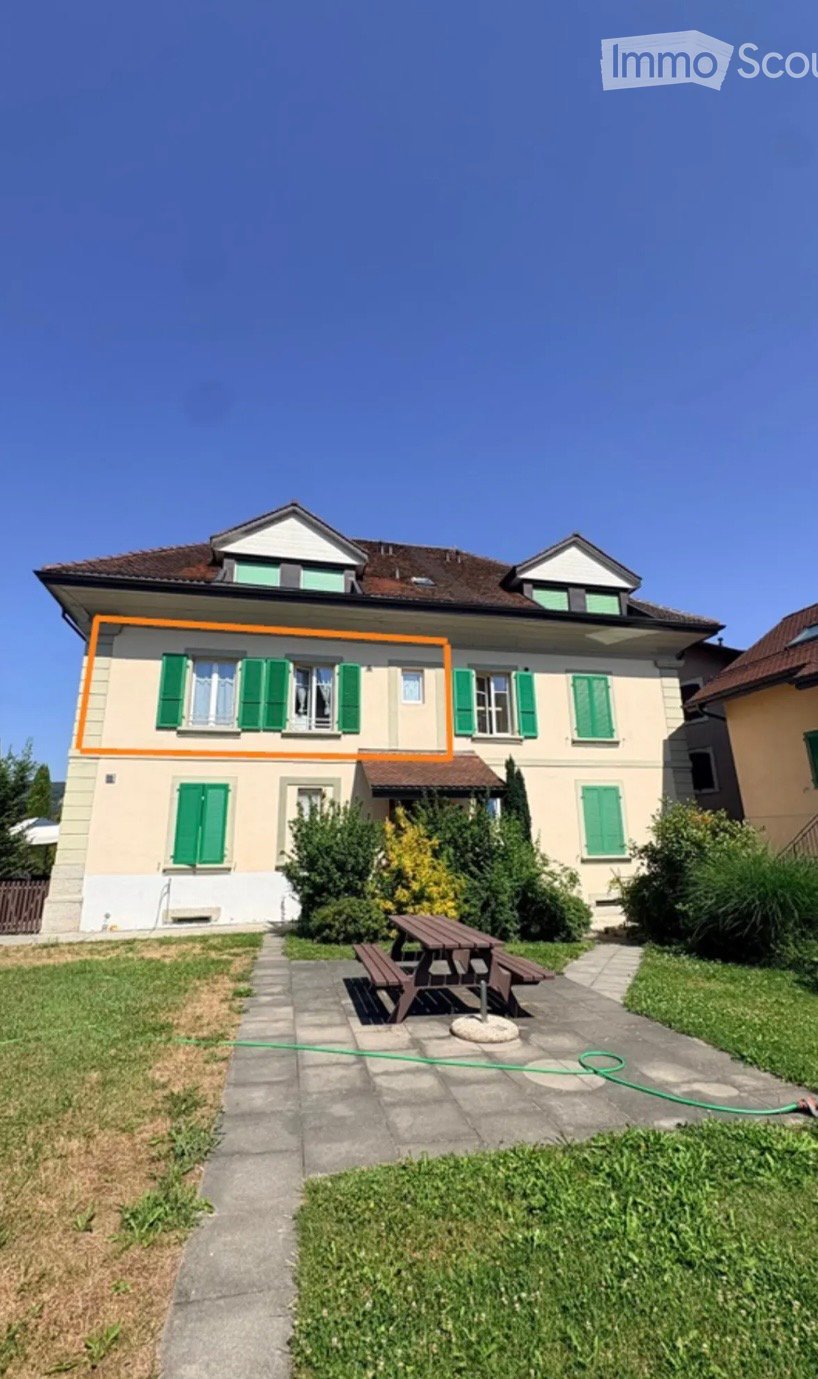 Two-story house with beige walls, green shutters, and a brown roof. The front yard includes a picnic table, benches, and a garden area.