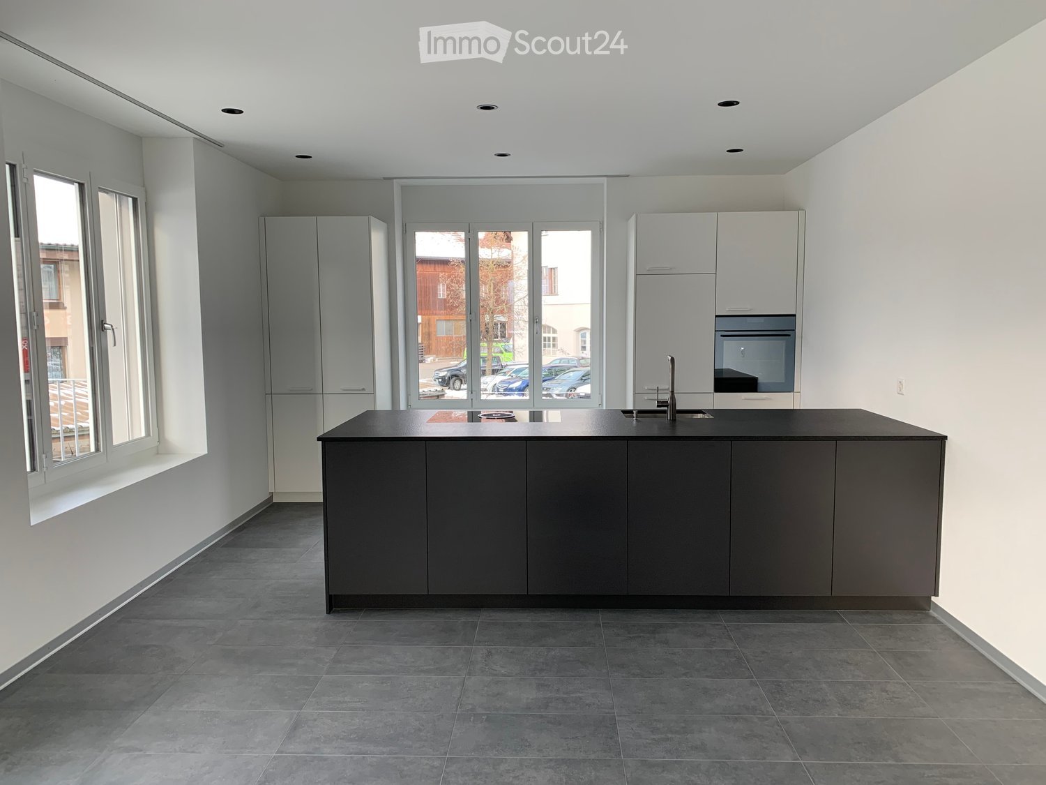 Modern kitchen with grey cabinets, a central island, and large windows overlooking a street with parked cars.