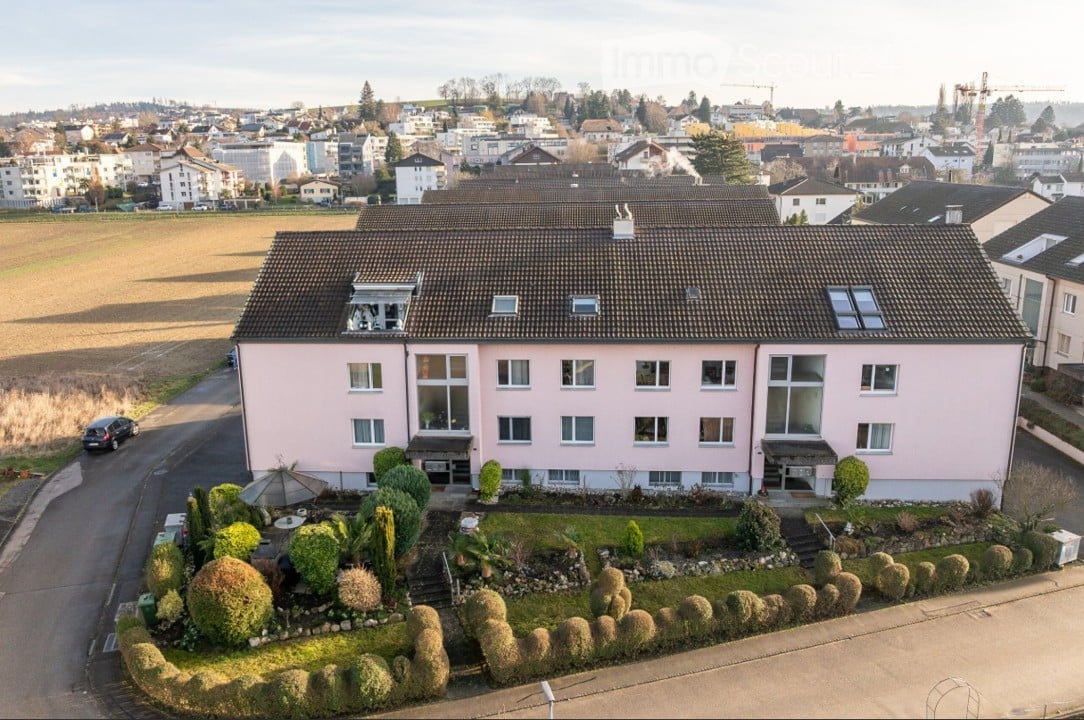 A two-story pink house with multiple windows and a well-maintained garden in front, situated in a residential neighborhood with other houses and a field in the background.