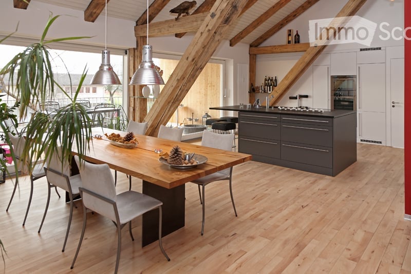 Open-plan kitchen and dining area with light wooden flooring, exposed wooden beams, modern kitchen island with cabinets, dining table with chairs, pendant lights, and decorative bowl on the table.