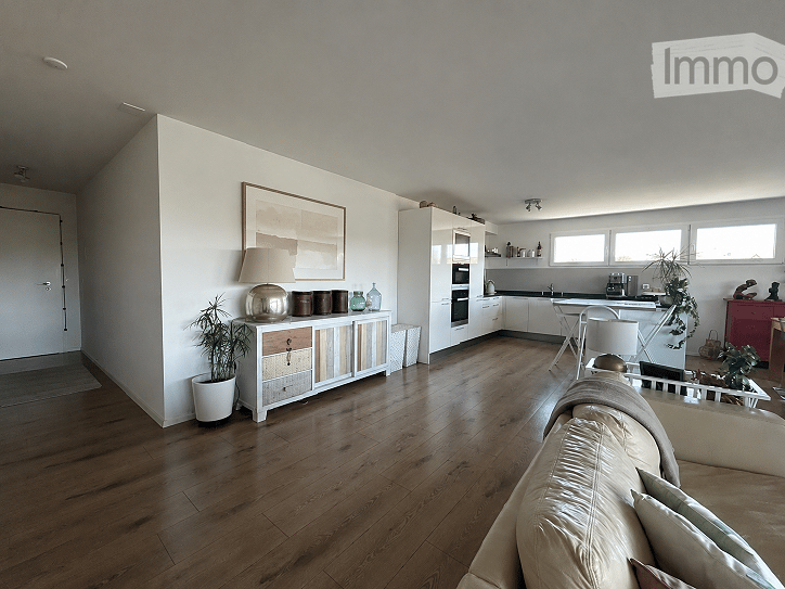 Open-plan living and dining area with hardwood flooring, white walls, a cream sofa, sideboard with decorative items, potted plants, and a modern kitchen area with cabinets and appliances.