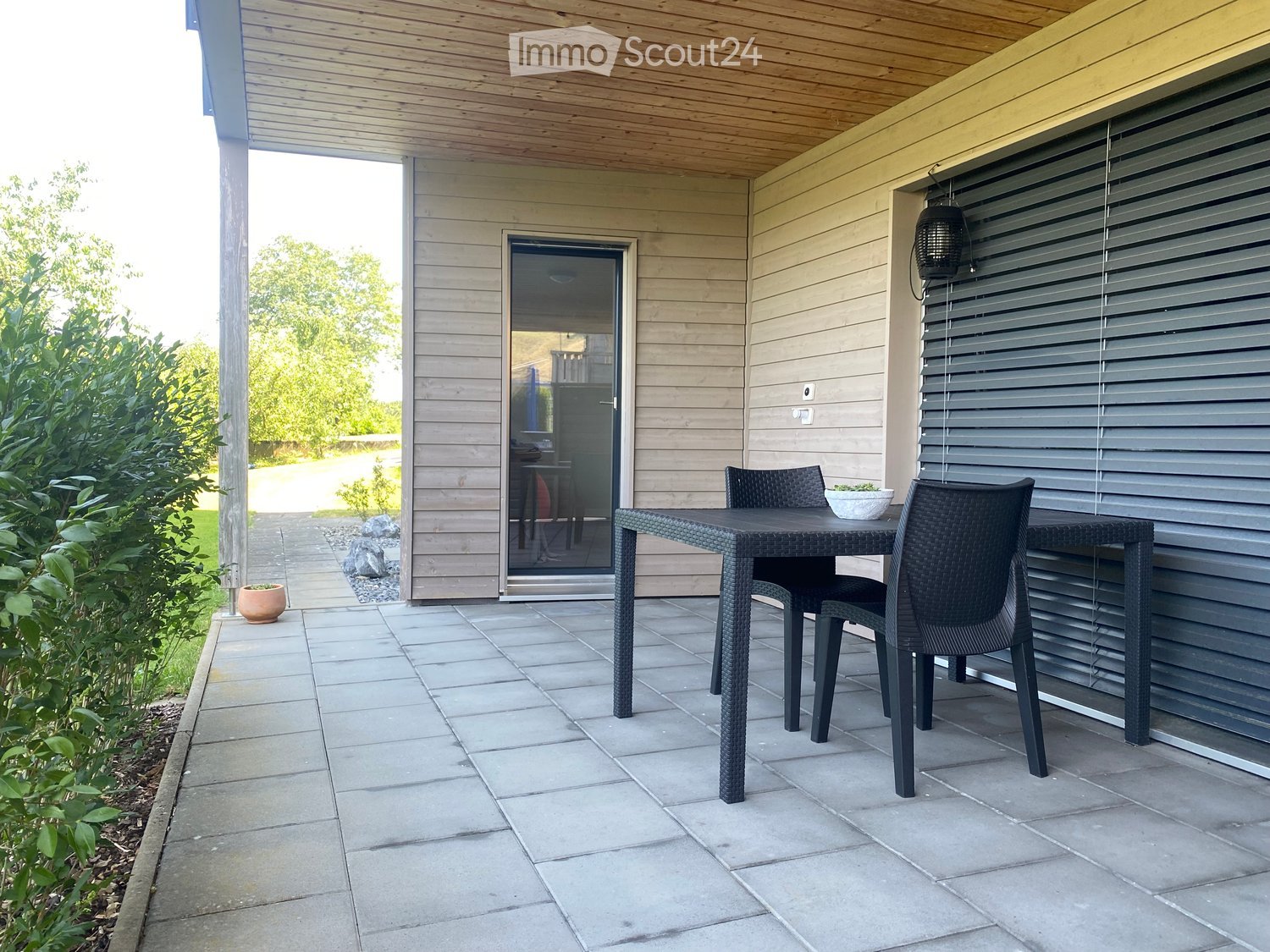 Outdoor patio with a wooden ceiling, tiled floor, and a black wicker dining set including a table and chairs. A sliding glass door leads to the interior, and there are potted plants and greenery nearby.